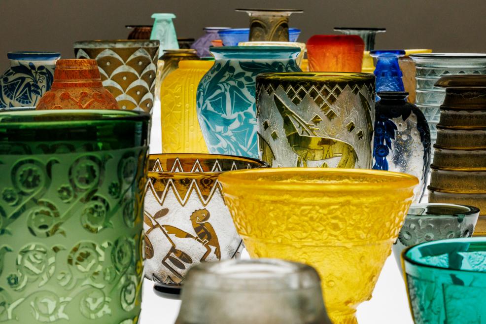 Krāsaini, dekoratīvi stikla trauki ar dažādiem gravējumiem un ornamentiem, cieši izvietoti uz apgaismotas virsmas.