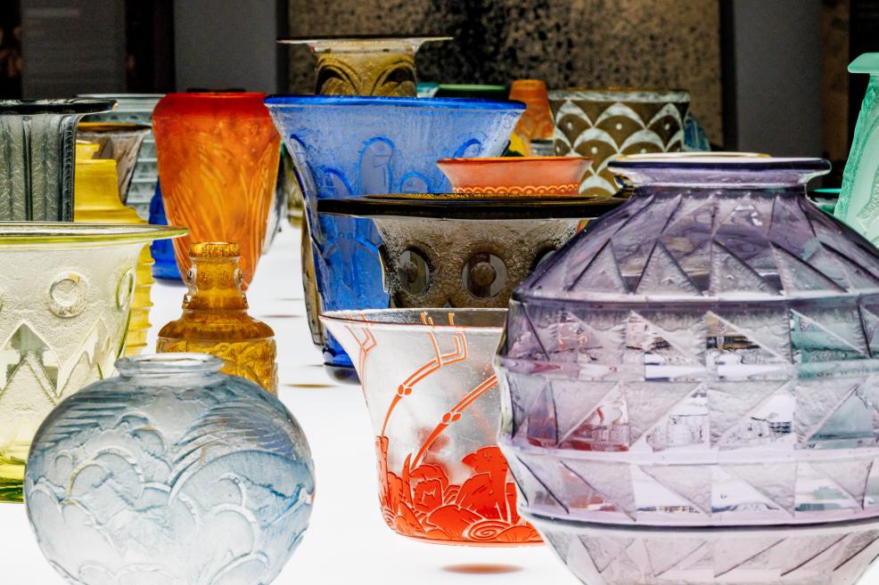 Dažādu formu un krāsu stikla trauki ar gravējumiem un reljefiem rakstiem, izkārtoti uz apgaismotas virsmas tuvplānā.