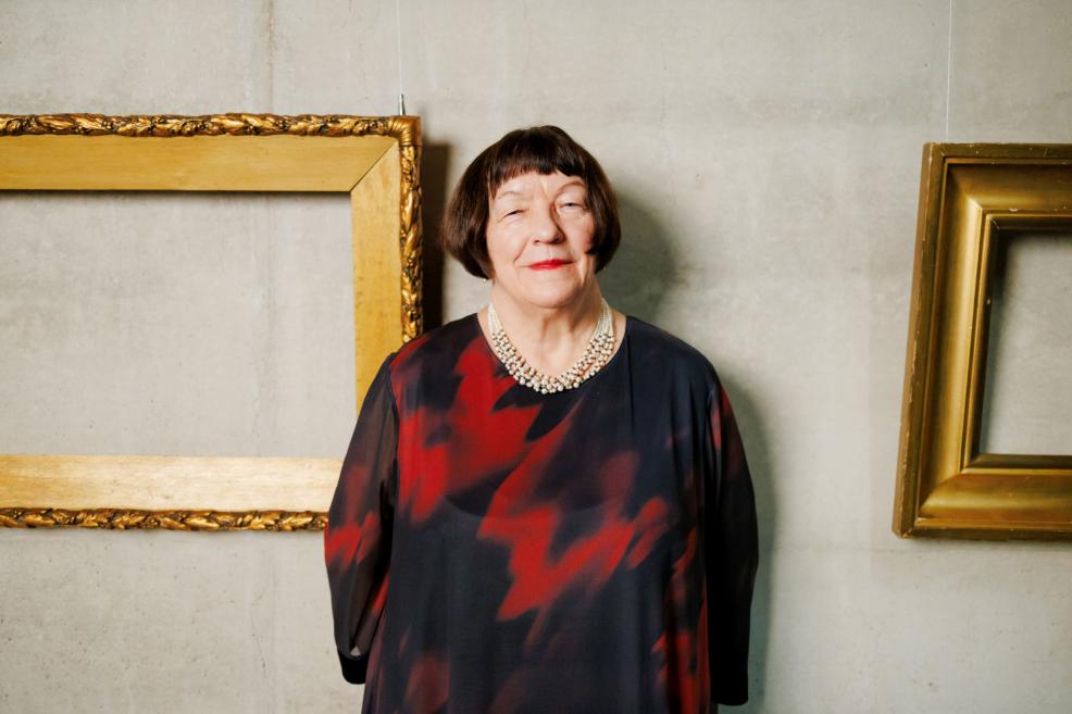 Sievietes portrets, stāvot pie sienas starp diviem tukšiem zeltītiem rāmjiem, valkājot tumšu kleitu un pērļu kaklarotu.