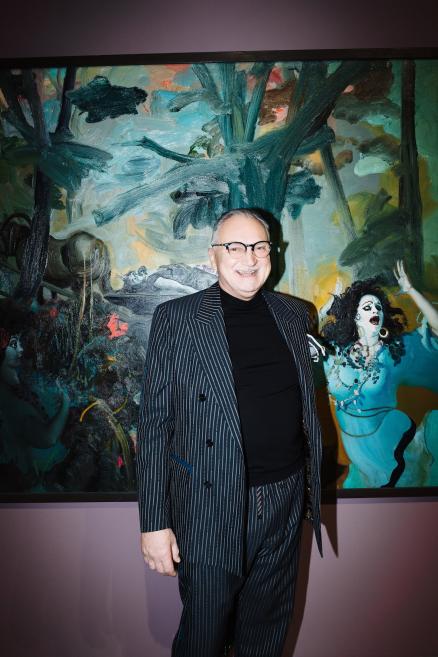 Vīrietis stāv pie lielas gleznas muzeja telpā, valkājot svītrainu uzvalku un brilles.