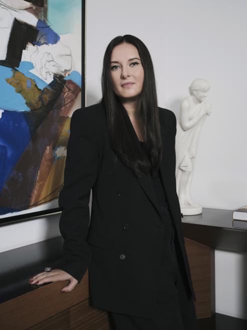 Sievietes portrets melnā uzvalkā, stāvot pie mēbeles interjerā ar abstraktu gleznu un skulptūru fonā.