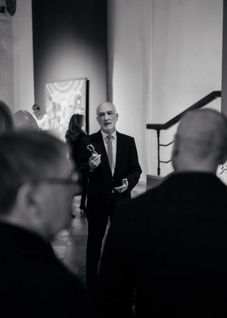 Vīrietis uzvalkā muzeja telpā uzrunā cilvēku grupu, rokā turot nelielu priekšmetu.