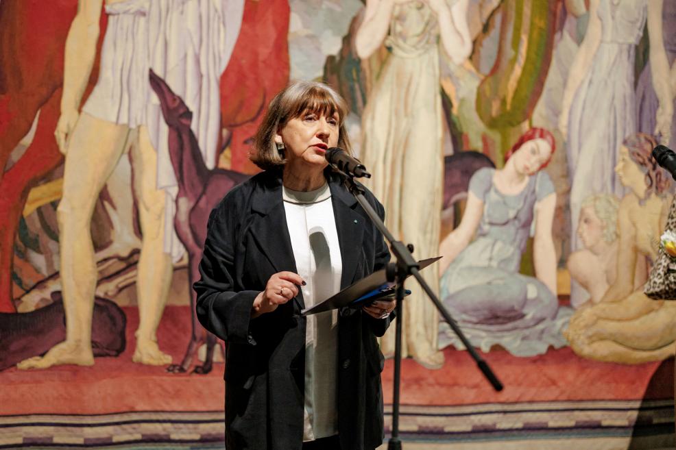 Sieviete runā mikrofonā muzeja telpā, rokā turot piezīmes, ar lielu figurālu gleznu fonā.