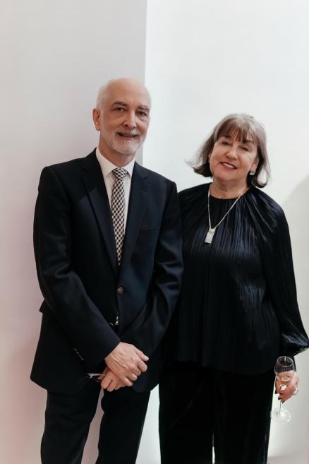 Vīrietis un sieviete formālā apģērbā stāv blakus, sievietei rokā vīna glāze, uz gaiša fona.