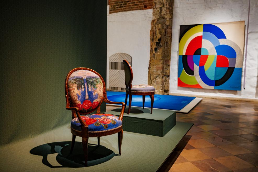 Muzeja ekspozīcijā izstādīti dekoratīvi krēsli ar rakstainu polsterējumu, fonā redzams krāsains ģeometrisks mākslas darbs.