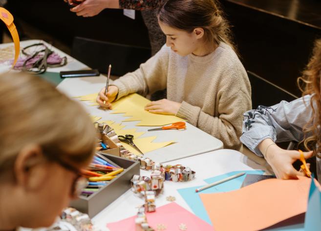 Bērni pie galda izgriež un zīmē krāsainu papīru radošā darbnīcā, izmantojot šķēres un zīmuļus.