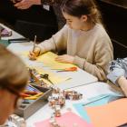 Bērni pie galda izgriež un zīmē krāsainu papīru radošā darbnīcā, izmantojot šķēres un zīmuļus.