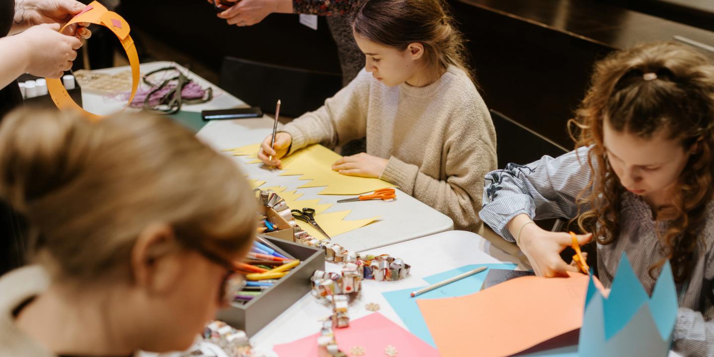 Bērni pie galda izgriež un zīmē krāsainu papīru radošā darbnīcā, izmantojot šķēres un zīmuļus.