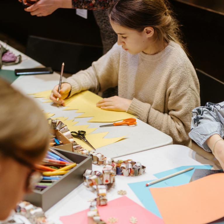 Bērni pie galda izgriež un zīmē krāsainu papīru radošā darbnīcā, izmantojot šķēres un zīmuļus.