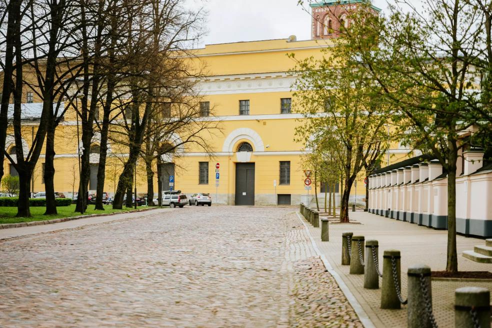 The wide façade of Riga Arsenāls with an entrance and cobblestone square in the foreground.
