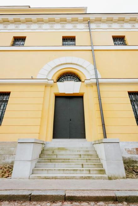 The main entrance of Riga Arsenāls with stairs and arched doors in the yellow façade.