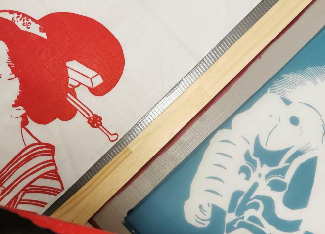 Auduma apdrukas darbnīcā redzami sietspiedes rāmji ar japāņu ukiyo-e stilā veidotiem portretiem sarkanā un zilā krāsā, ar uzklātu krāsu un koka līstēm darba procesā.