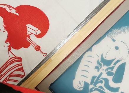 Auduma apdrukas darbnīcā redzami sietspiedes rāmji ar japāņu ukiyo-e stilā veidotiem portretiem sarkanā un zilā krāsā, ar uzklātu krāsu un koka līstēm darba procesā.