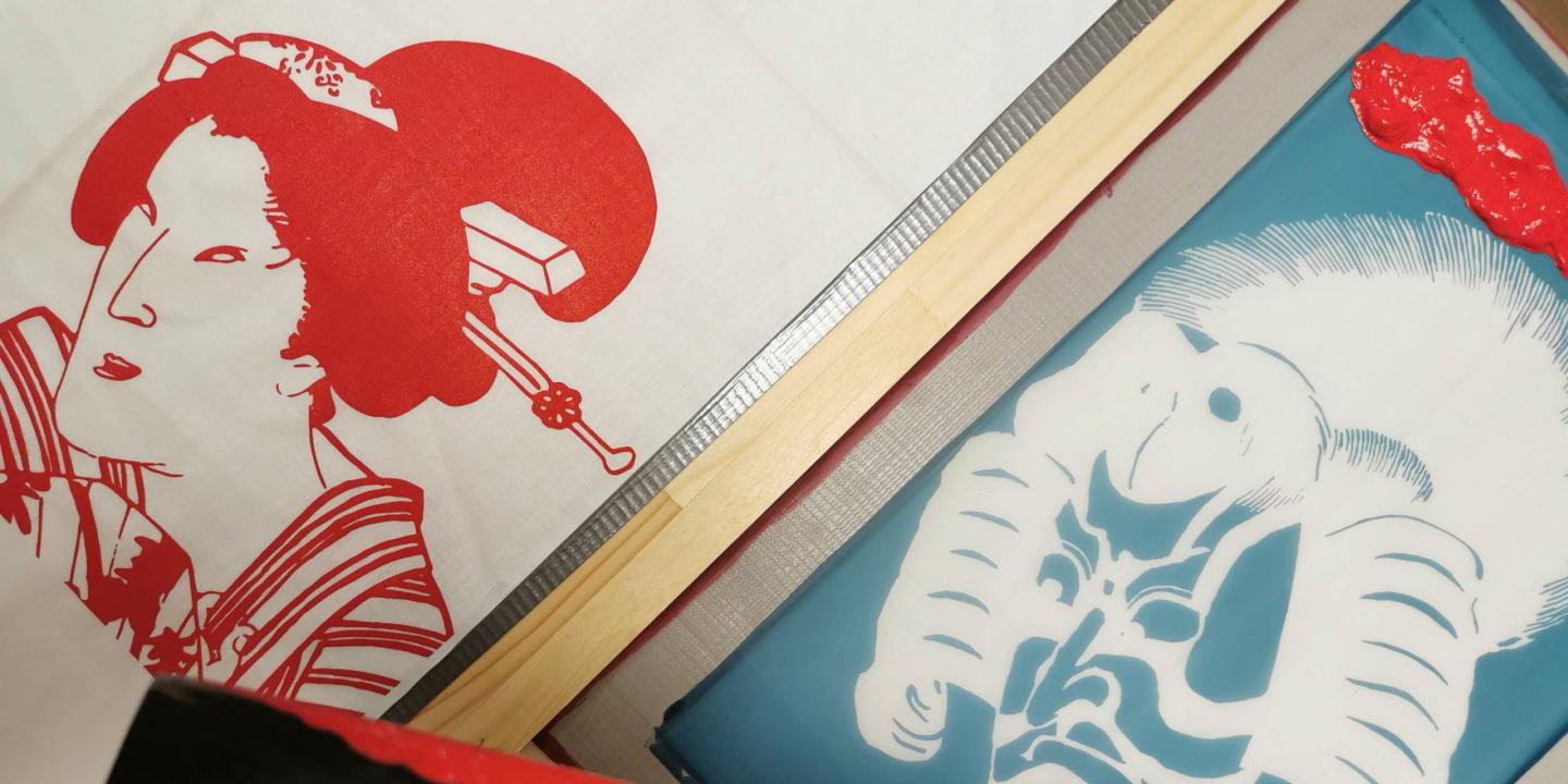 Auduma apdrukas darbnīcā redzami sietspiedes rāmji ar japāņu ukiyo-e stilā veidotiem portretiem sarkanā un zilā krāsā, ar uzklātu krāsu un koka līstēm darba procesā.