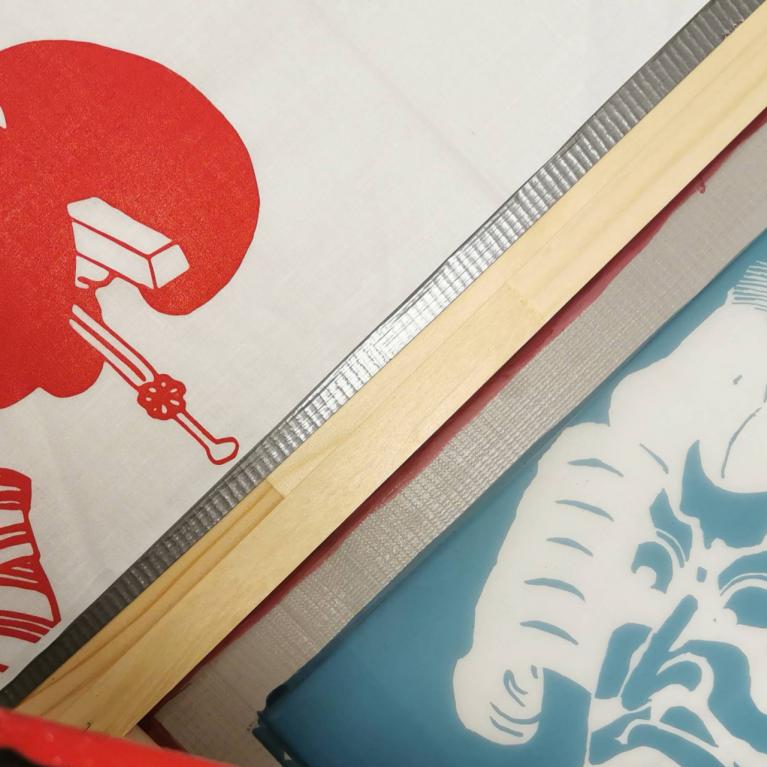 Auduma apdrukas darbnīcā redzami sietspiedes rāmji ar japāņu ukiyo-e stilā veidotiem portretiem sarkanā un zilā krāsā, ar uzklātu krāsu un koka līstēm darba procesā.