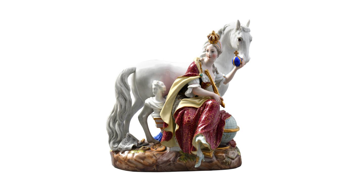Porcelāna figūriņa ar kronētu sievieti krāsainā tērpā, kura sēž pie balta zirga un tur zilas lodes ar krustu, uz dekoratīvas pamatnes.