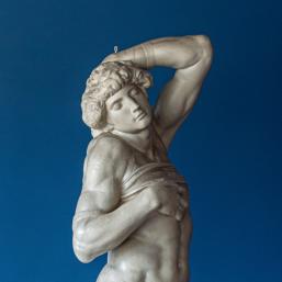 Marble sculpture of a male figure in a dramatic pose, with one arm raised and head tilted, set against a blue background.