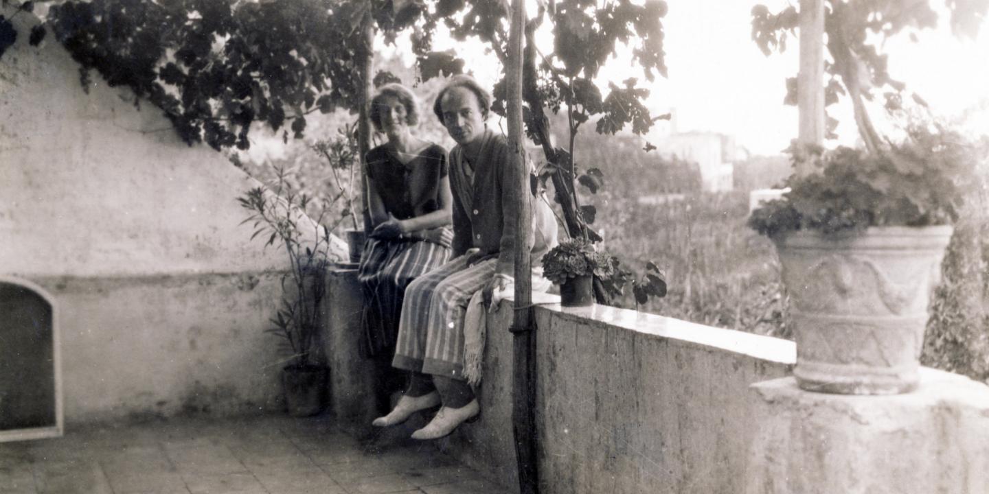 Vēsturiska melnbalta fotogrāfija ar diviem cilvēkiem, kas sēž terasē zem vīnogulāju zariem.