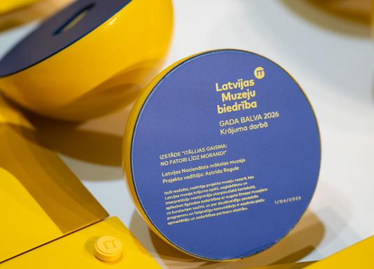 Uz galda izvietotas Latvijas Muzeju biedrības Gada balvas 2026 trofejas dzeltenā un zilā krāsā; priekšplānā redzama balva par krājuma darbu izstādei “Itālijas gaisma: no Fatori līdz Morandi”.