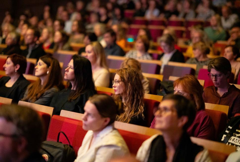 Auditorijā sēdoši klausītāji uzmanīgi seko pasākuma norisei, aizpildot vairākas rindas ar sarkaniem krēsliem.