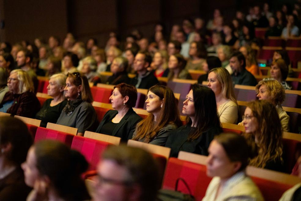 Auditorijā sēdoši pasākuma apmeklētāji koncentrēti klausās priekšnesumu vai prezentāciju, aizpildot vairākas rindas ar sarkaniem krēsliem.