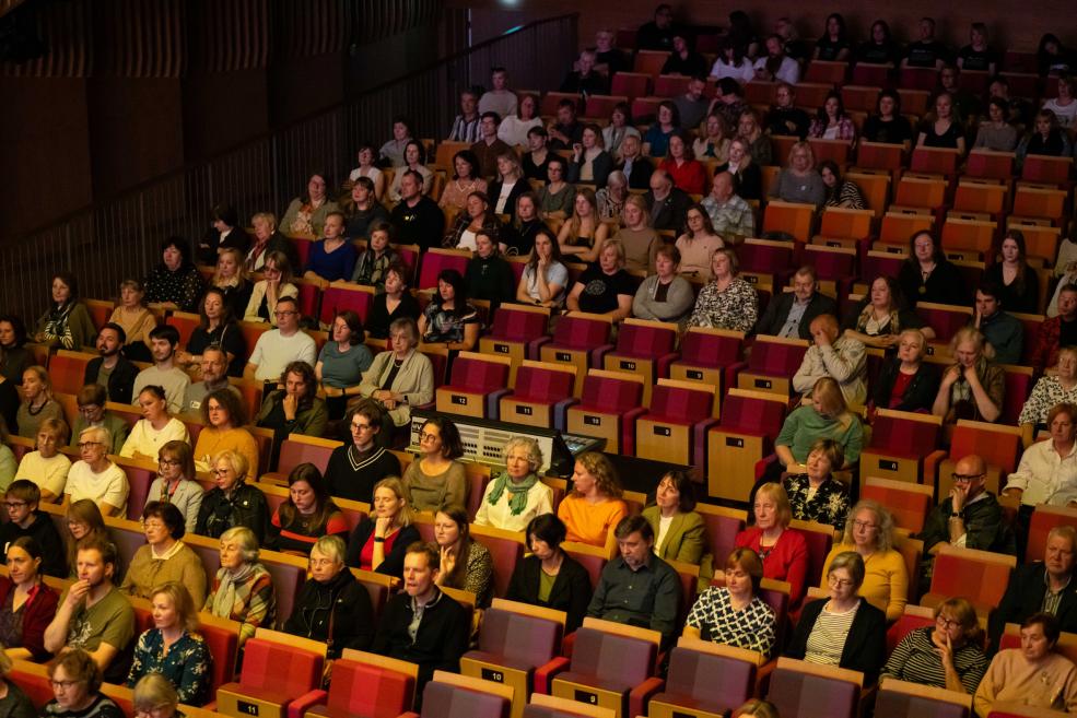 Pilna auditorija ar desmitiem klausītāju sēž amfiteātra tipa zālē un uzmanīgi seko pasākuma norisei.