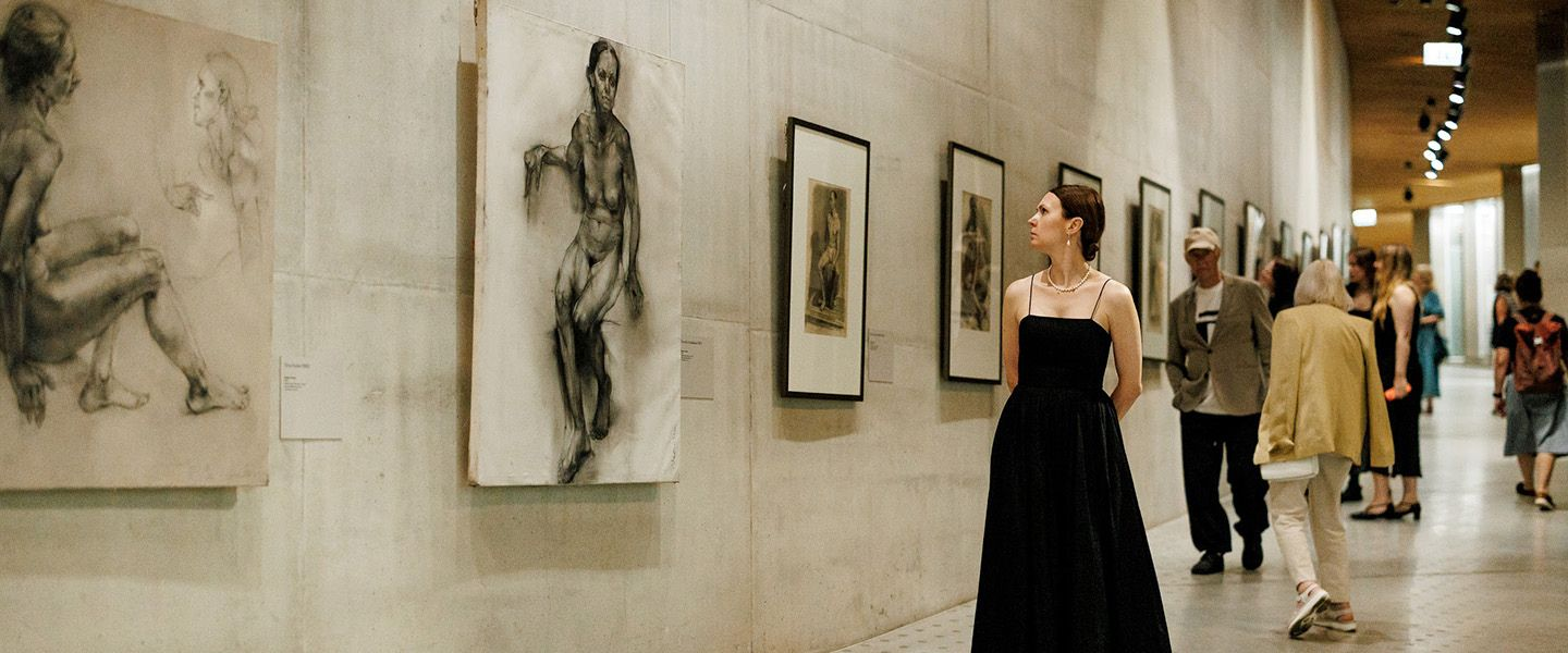 A woman in a black dress looks at a figurative drawing in an art museum gallery where several framed artworks are displayed on the wall.