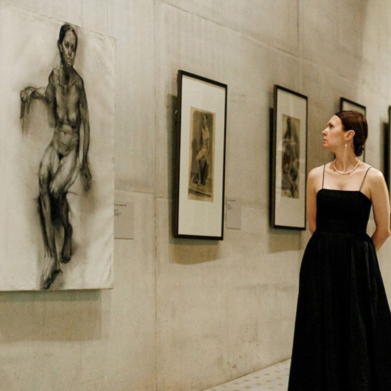 A woman in a black dress looks at a figurative drawing in an art museum gallery where several framed artworks are displayed on the wall.