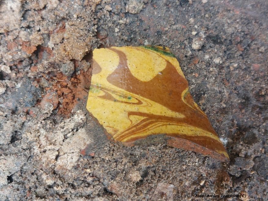 Zemē atrasts dekorētas keramikas fragments ar dzeltenas un brūnas glazūras viļņveida rakstu, daļēji klāts ar smiltīm un augsni; redzamas nolietojuma un laika radītas pēdas.