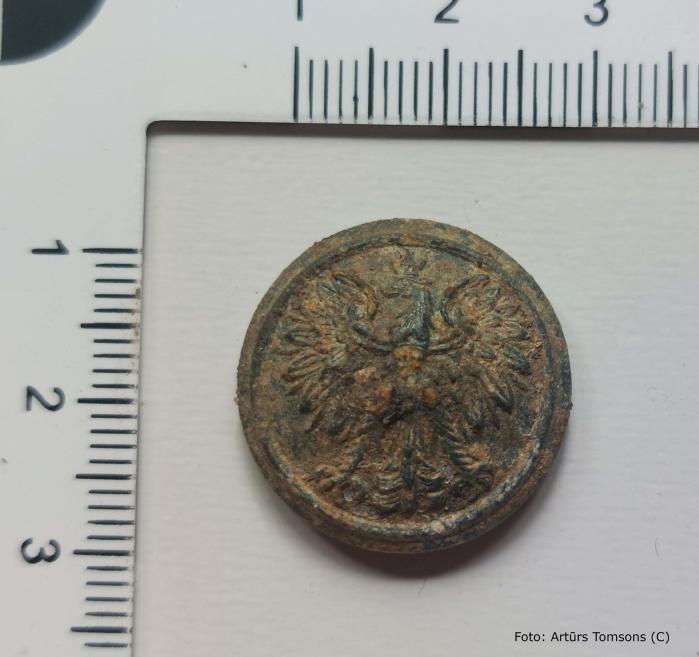 Apaļš metāla priekšmets ar reljefu dekoru (auglim vai ziedam līdzīgu motīvu), stipri oksidējies un korodējis, fotografēts uz neitrāla fona ar lineālu mēroga noteikšanai.