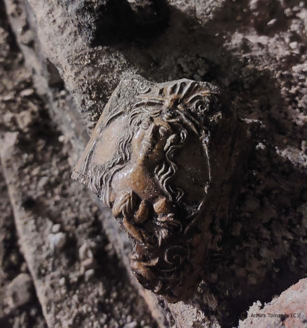 Zemē daļēji ieaudzis metāla fragments ar reljefā veidotu cilvēka sejas profilu, redzamas matu cirtas un sejas vaibsti; priekšmets klāts ar netīrumiem un patīnu, fotografēts tuvplānā.