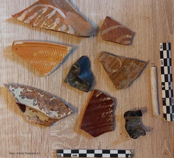 Uz koka virsmas izkārtoti dažādi arheoloģiski atradumi – keramikas lausku fragmenti ar glazējumu un rievotu faktūru, tumšs akmens gabals un neliels cilindrisks priekšmets; blakus redzamas mērlentes centimetru skalas izmēra noteikšanai.