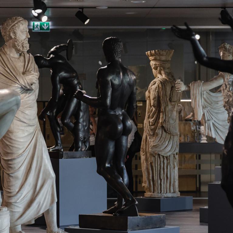 Muzeja zālē izvietotas klasiskas skulptūras – marmora un tumša metāla cilvēkfigūras dažādās pozās uz postamentiem.