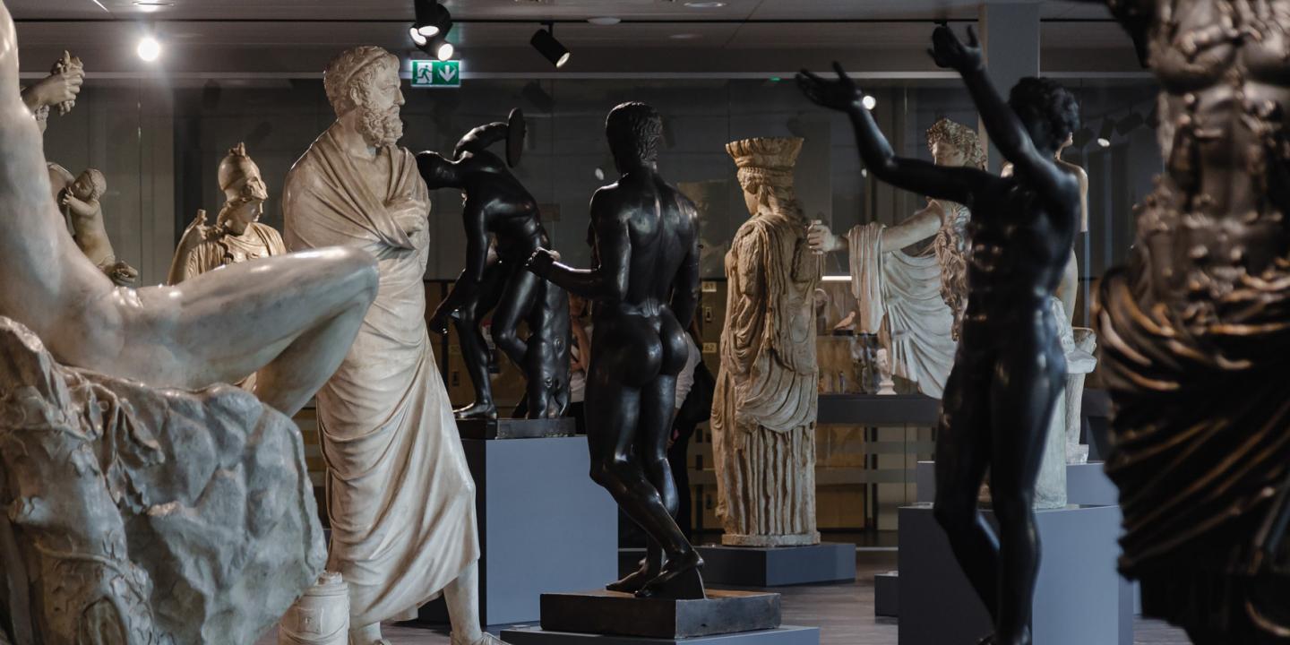 Muzeja zālē izvietotas klasiskas skulptūras – marmora un tumša metāla cilvēkfigūras dažādās pozās uz postamentiem.