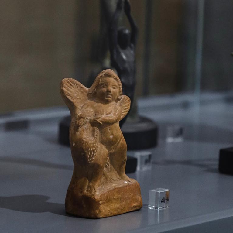 Vitrīnā izstādītas nelielas skulptūras, tostarp spārnota figūra un citas cilvēkveidīgas statuetes uz pamatnēm.
