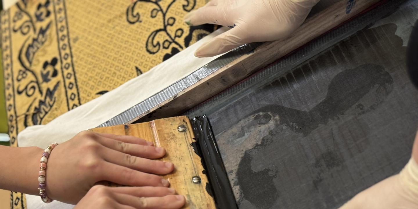 Tuvplānā redzamas rokas, kas ar sietspiedes rāmi un lāpstiņu klāj tinti uz auduma ar ornamentu.