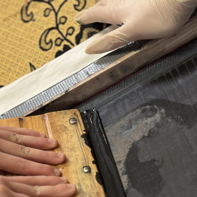 Tuvplānā redzamas rokas, kas ar sietspiedes rāmi un lāpstiņu klāj tinti uz auduma ar ornamentu.