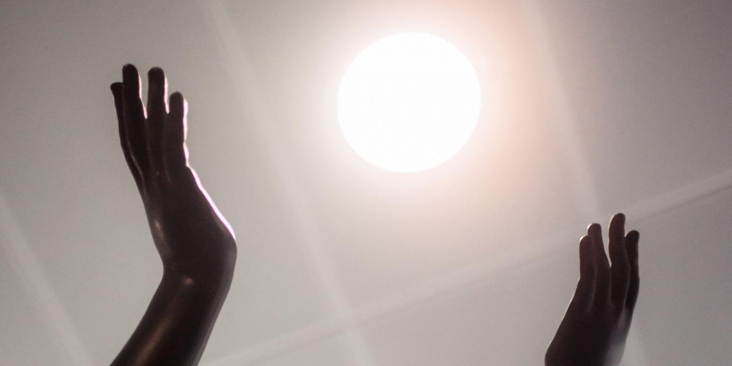 Tumšas skulptūras rokas paceltas pret spožu gaismas apli, kas atgādina sauli.