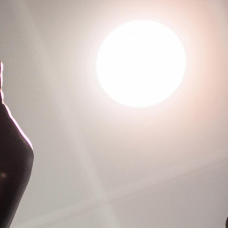 Tumšas skulptūras rokas paceltas pret spožu gaismas apli, kas atgādina sauli.