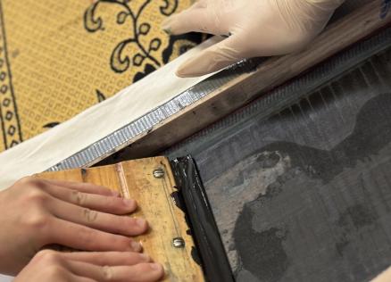 Tuvplānā redzamas rokas, kas ar sietspiedes rāmi un lāpstiņu klāj tinti uz auduma ar ornamentu.