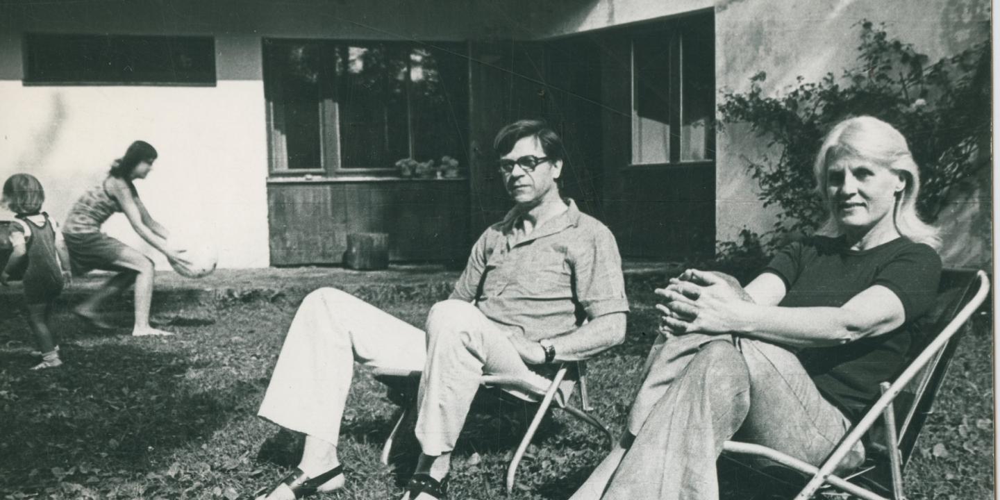 Melnbaltā fotogrāfijā dārzā uz krēsliem sēž divi pieaugušie, kamēr fonā bērni spēlējas pie mājas.