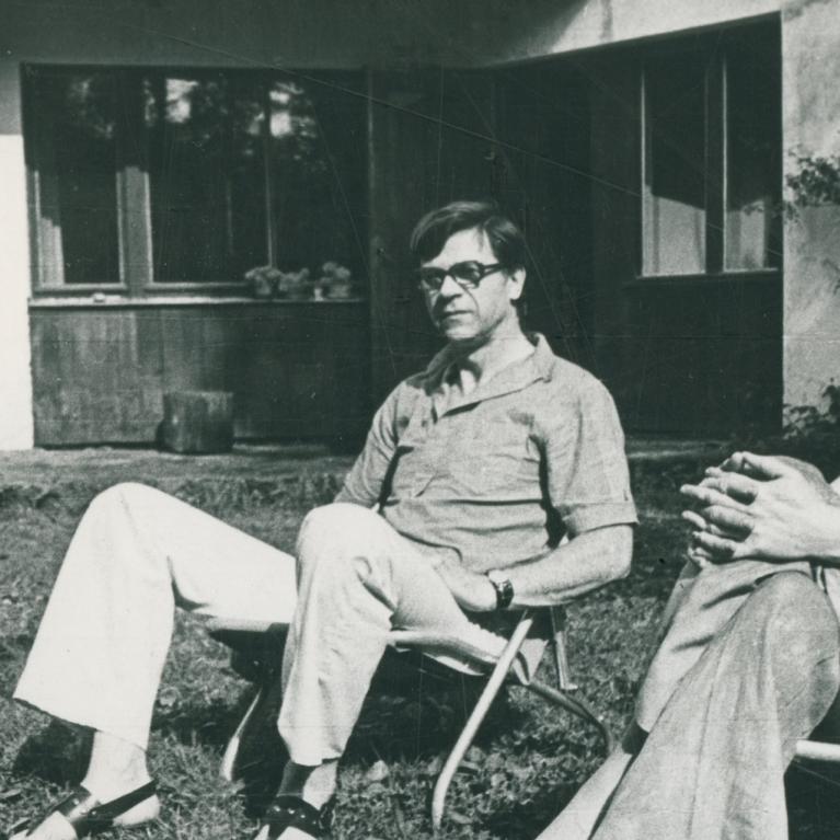 Melnbaltā fotogrāfijā dārzā uz krēsliem sēž divi pieaugušie, kamēr fonā bērni spēlējas pie mājas.