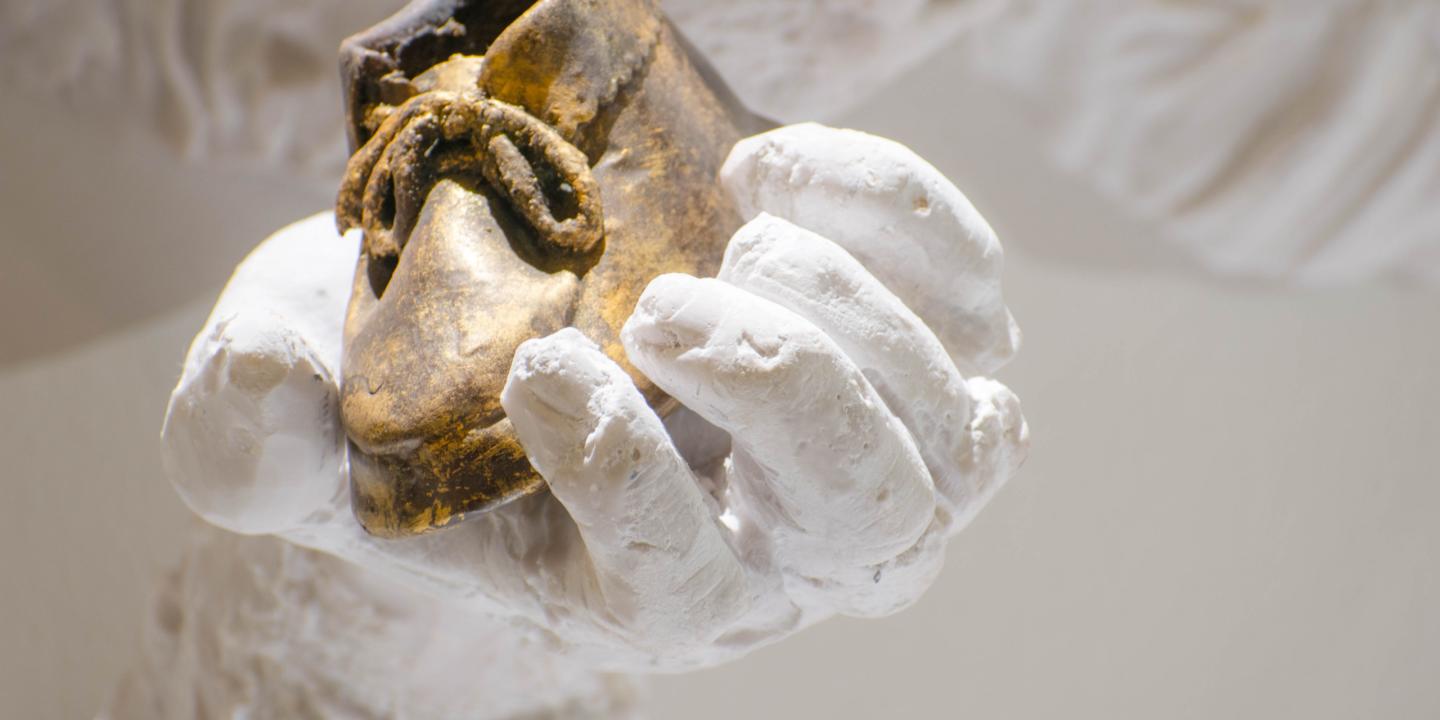 Baltas skulpturālas rokas tuvplānā tur nelielu zeltītu bērna kurpīti, izceļot kontrastu starp materiāliem un faktūrām.