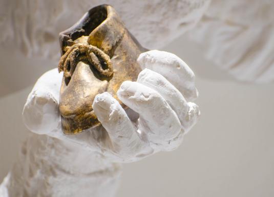 Baltas skulpturālas rokas tuvplānā tur nelielu zeltītu bērna kurpīti, izceļot kontrastu starp materiāliem un faktūrām.