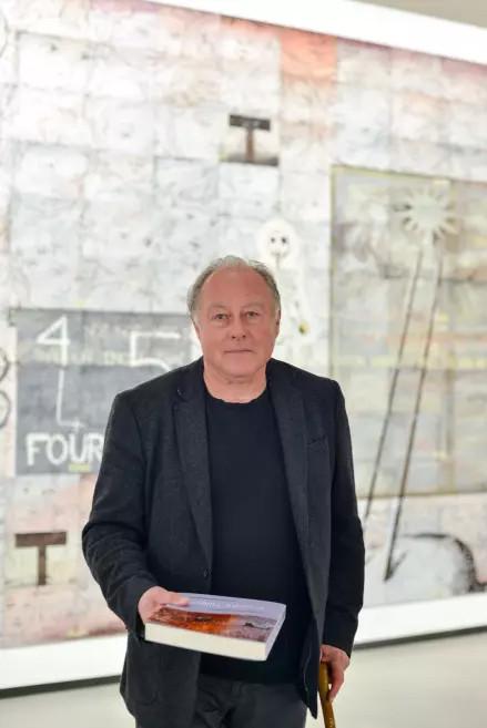 Portrait of the artist in the exhibition hall holding a catalogue in front of a monumental artwork.