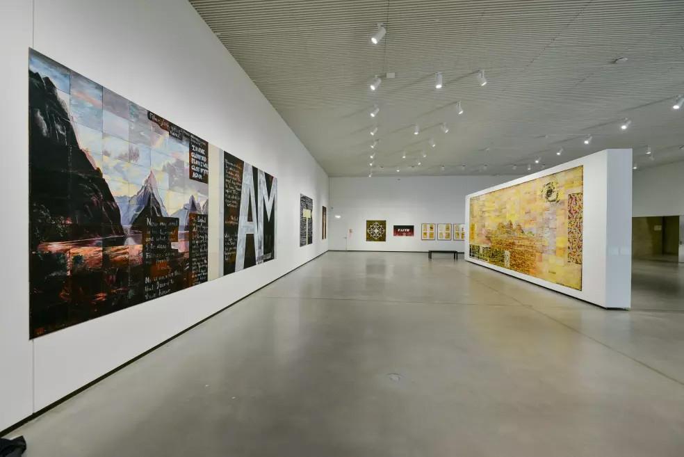 A spacious exhibition hall with large paintings on the walls, displaying texts, landscapes, and color compositions.