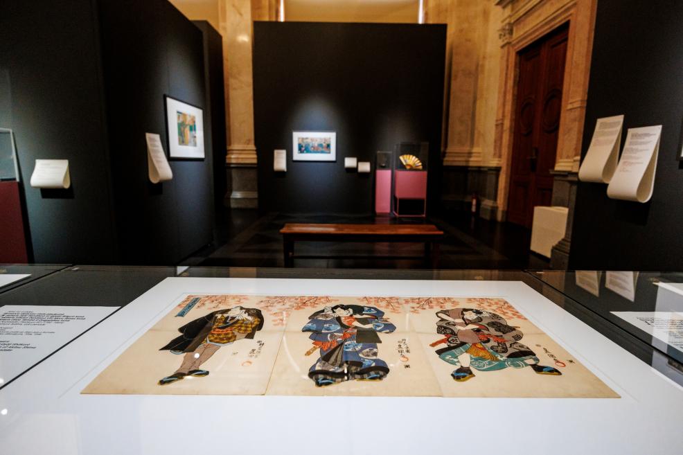 A view of a museum exhibition featuring a Japanese print displayed in a glass case, with other works shown on dark walls in the background.
