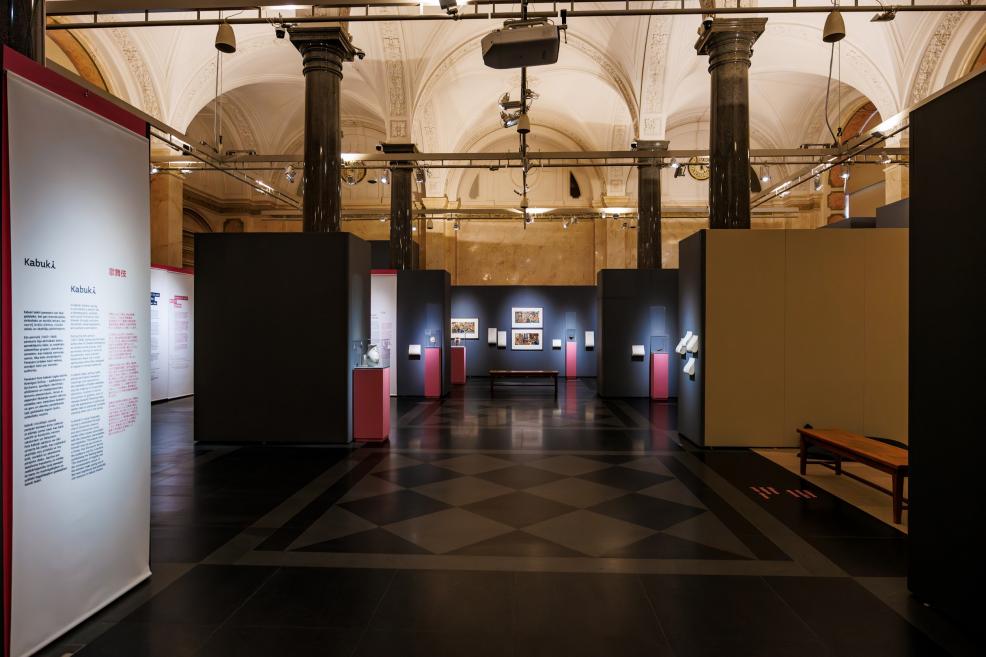 A general view of a museum exhibition with high ceilings and columns, featuring works and display cases of Japanese art set against dark walls.