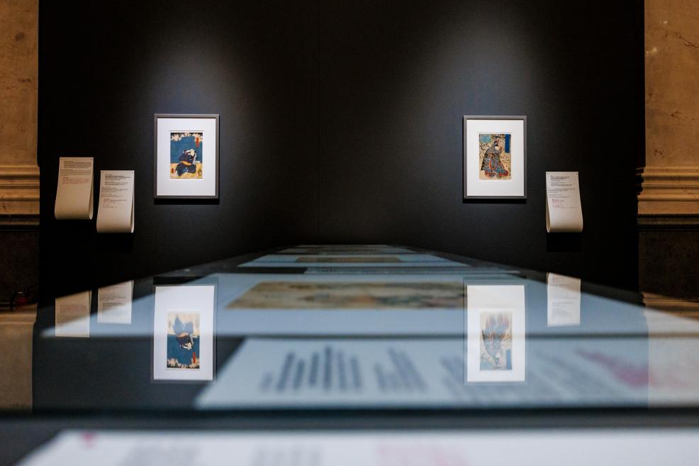 A view of a museum exhibition with two framed Japanese prints on a dark wall and the reflection of a display case in the foreground.