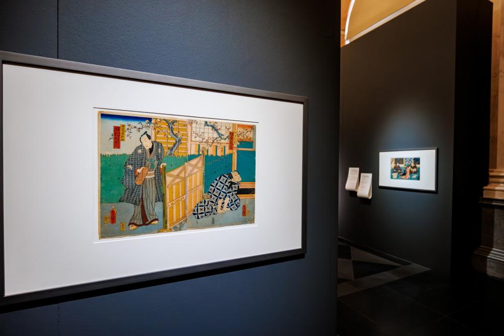 A framed Japanese color print depicting figures in an interior is displayed on a dark wall in a museum exhibition.